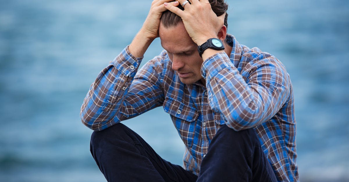 A man in a plaid shirt sits by the water looking distressed, symbolizing stress. headache.