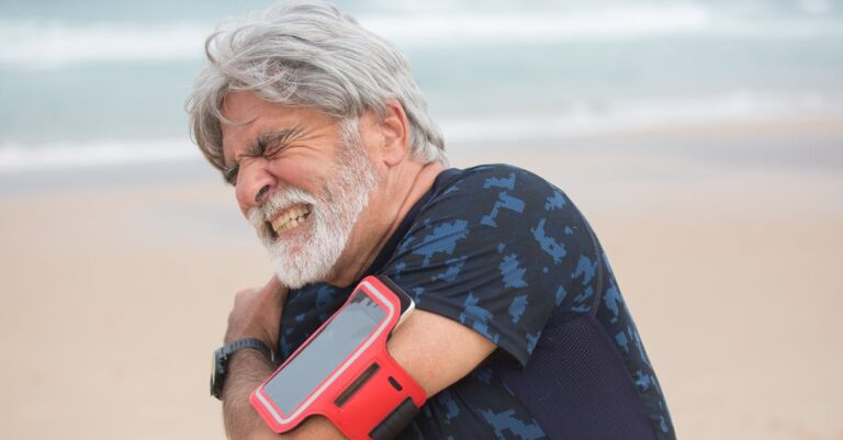 Elderly man grimacing from shoulder pain during seaside walk, showcasing health issue outdoors.