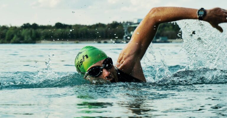 A male swimmer in a green cap and goggles competing in an open water race. The repetitive strain of swimming can result in scar tissue formation in the rotator cuff