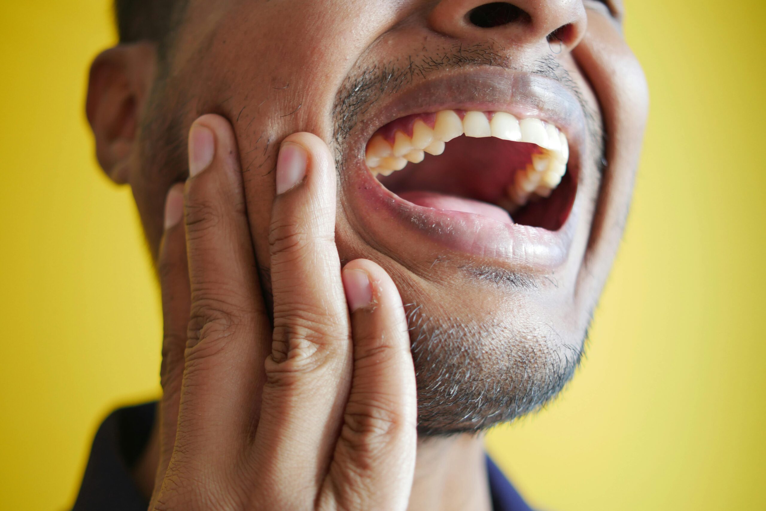 Close-up of a man holding his cheek, displaying a wide open mouth against a yellow background. See our TMJ chiropractor for jaw pain.