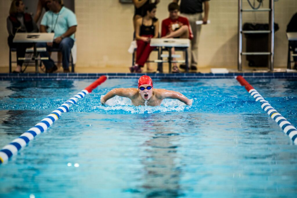 🟦 Swim Stronger, Recover Smarter: Chiropractic for Swimmers — How Chiropractic Can Elevate Their Stroke 3 A young swimmer in a red cap competes in a butterfly stroke during an indoor pool race. Chiropractic for swimmers.