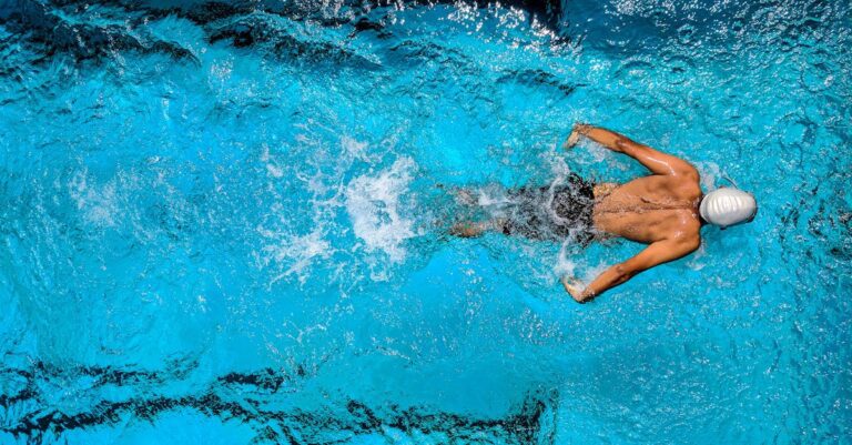 Top view of a swimmer wearing a cap, performing a front crawl stroke in a clear blue swimming pool. Chiropractic for swimmers.