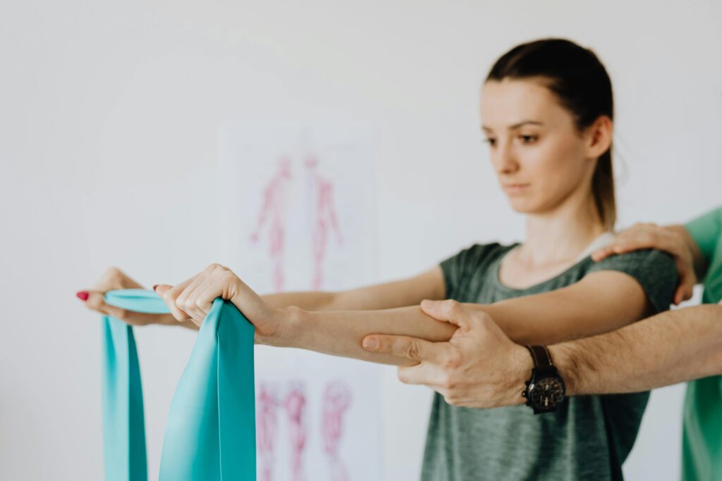 Slim young female in casual wear standing with reached arms and elastic tape during medical checkup by crop unrecognizable chiropractor in wristwatch