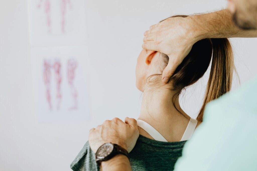 Vancouver Chiropractor in Yaletown treating a patient. Back view of crop faceless bearded orthopedist in wristwatch checking up neck of anonymous lady while standing in front of wall with paper drawings representing body anatomy